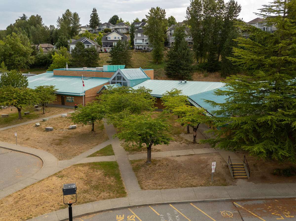 Exterior image of the entrance into an elementary school. Canadian flag pole out front with neighbourhood homes in the background. Building has lots of green trees surrounding it.