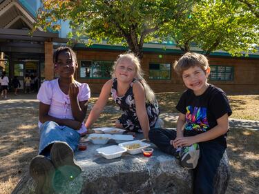Three elementary students sitting on a rock, eating french fries.