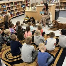 Group of students sitting on carpet in the library listening to an adult read a story, as they have two dogs sitting next to them.